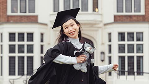 Xu wearing a graduation gown and cap holding her degree scroll outside Hazlerigg Building on 麻豆視頻_麻豆直播_麻豆传媒官网 campus. She is smiling and she is also holding a grey stuffed teddy bear which is also wearing a black cap and gown.